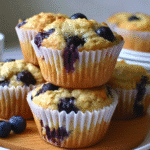 Freshly baked blueberry oatmeal muffins in white paper liners, stacked on a plate and surrounded by fresh blueberries.